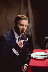 handsome man in suit sitting at table and lighting up cigar in restaurant