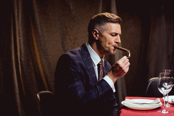 handsome man in suit sitting at table and lighting up cigar in restaurant