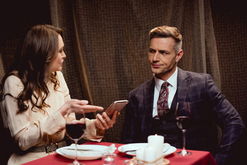 woman holding smartphone and arguing with dissatisfied man during romantic date in restaurant
