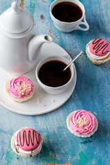 Vanilla cupcakes with pink cream, a coffee pot and a cup of coffee on an old wooden table. 