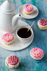 Vanilla cupcakes with pink cream, a coffee pot and a cup of coffee on an old wooden table. 