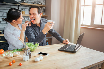 Fototapeta premium Cheerful people sit together in room. Woman feed guy with tometo. He keep moouth opened. Man hold white cup.