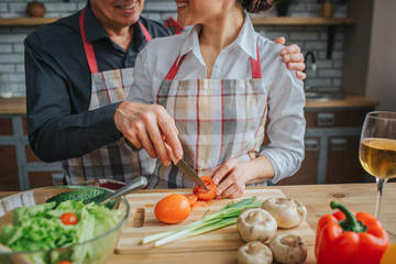Cut view of man and woman stand together and cooking at table in kitchen. He has knife and cut tomato. She hold red vegetable with hands. They look at each other and smile.