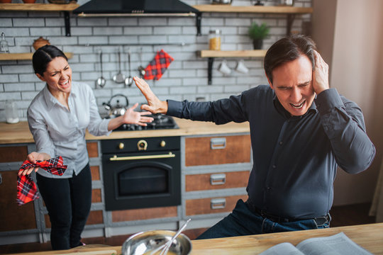 Couple On Kitchen. Man Sit At Table And Close His Ears. He Show Hand. Woman Try To Talk To Him. They Argue.