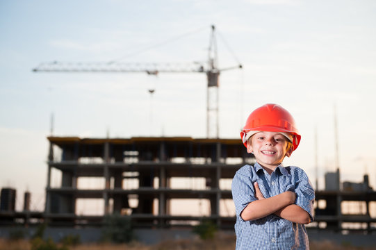 handsome happy smiling little caucasian kid in orange helmet and blue shirt standing on construction site with crane