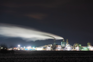 A night view of an industrial unit, a wood-processing plant with fuming pipes and a high-illuminated area on the background of a snow-covered field and a bicycle with a red light.