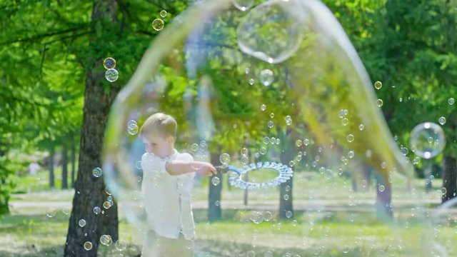 Slow motion of adorable little blonde boy spinning with wand and blowing soap bubbles around while playing with friends on green lawn in the park at summer day