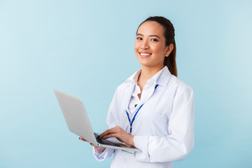 Young woman doctor posing isolated over blue wall background using laptop computer.