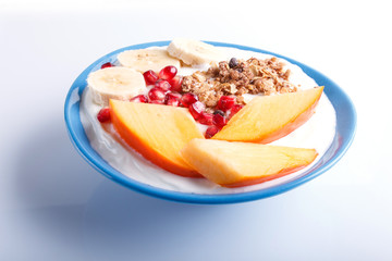 A blue plate with greek yogurt, granola, persimmon, banana, pomegranate isolated on white.