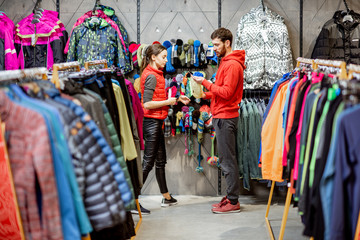 Man and woman looking for winter clothes standing together in the sports shop with colorful jackets...