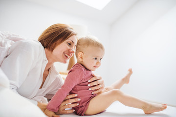 A young mother with little daughter sitting indoors on bed in the morning, playing.