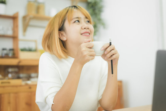 Beautiful young smiling woman working on laptop while enjoying drinking warm coffee sitting in a living room at home. Enjoying time at home. Asian business woman working in her home office.