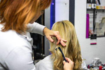 Professional hairdresser using comb and her fingers for making curls on beautiful blonde hair. Client sitting on the chair