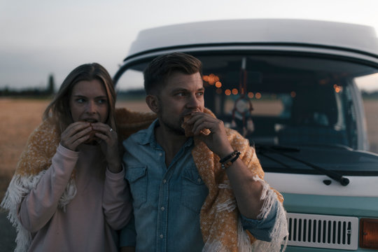 Couple wrapped in a blanket at camper van having a snack in rural landscape