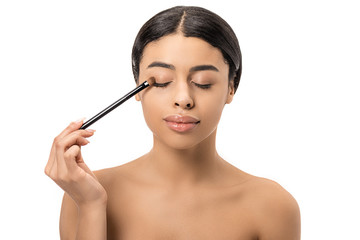 Fototapeta premium attractive brunette african american girl with closed eyes applying eyeshadow with cosmetic brush isolated on white