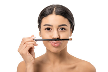 attractive young african american woman holding makeup brush on lips and looking at camera isolated on white