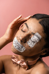 Naklejka premium beautiful sensual african american woman with paint on face and bow tie touching hair isolated on pink