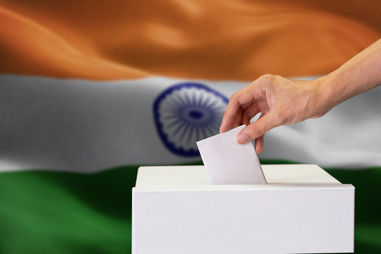 Close-up of human hand casting and inserting a vote and choosing and making a decision what he wants in polling box with India flag blended in background.