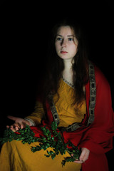 Young white Caucasian woman with long red hair in the form of a Greek goddess is sitting on a black background in a yellow dress and a red raincoat with a sprig of olives in her hands - obrazy, fototapety, plakaty