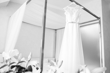 Beautiful bride's white wedding dress hangs near the bed in a hotel room with flowers at the bottom. Bridal morning before ceremony. Black and white photo.