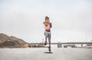 Strong athletic African American Black woman wearing long black and white printed tights and a pink sports bra is sprinting or running on a hot road in a dusty rocky desert background  