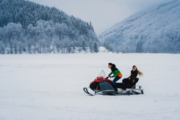 Couple riding on a snowmobile on the frozen lake in the mountains with the scenic view. Pine trees covered with snow. Side view