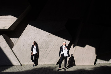 Couple of woman in classic suits and with red lipstick are posing on concrete background, smiling together, hanging 