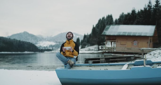 A happy tourist in the amazing nature place with a snowy lake and mountain with a big forest in the background sitting in the boat and playing with a tablet , charismatic tourist spending a nice time