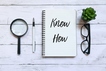 Top view of magnifying glass,glasses,plant,pen and notebook written with Know How on white wooden...
