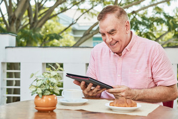 Happy aged man eating breakfast and reading news on tablet computer