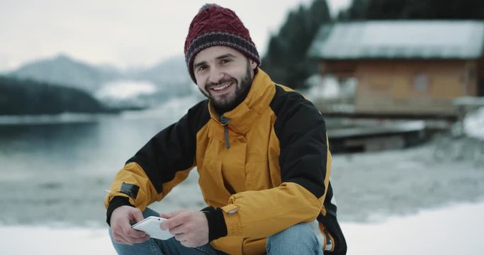 In a winter day charismatic young tourist in a orange jacket and red hat happy sitting beside the lake shore in a blue boat closeup smiling large looking straight to the camera and showing a big