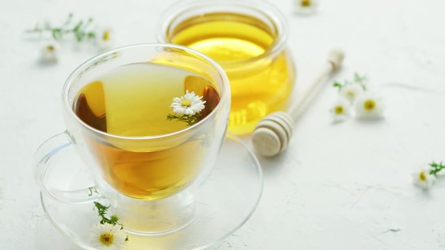 Transparent glass cup with herbal camomile tea and fresh flowers on table with jar of honey and wood stick