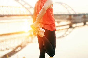 Close up of Caucasian woman stretching leg. In background river, bridge and morning sun.