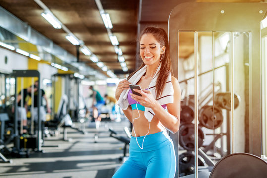 Picture of cute smiling sporty girl in gym preparing her self for the training. Choosing music on her telephone .Posing in front of the camera and looking at it.