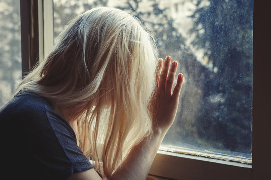Blonde haired woman leaning against window, looking out, sad, depressed and lonely with unbrushed long hair, face not visible 