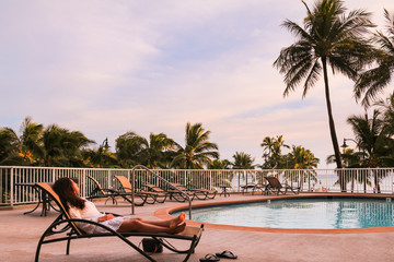 Young asian woman reclines back on a sunbed next to a swimming pool.