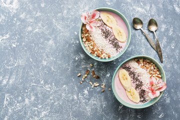 Pink yogurt with almonds, banana, chia seeds and coconut flakes in blue bowls on a gray background decorated with flowers. Healthy breakfast. Top view, copy space.