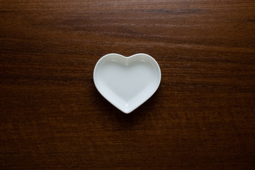 Empty plate in the shape of a heart on a wooden table in the center of the frame. Ceramic glossy dishes on a dark nutty background. Copy space. Concept of Valentine's Day or wedding romantic theme.