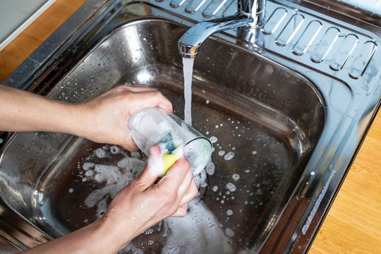 A human hand washes a glass with a sponge and detergent under clean running water, in the sink.