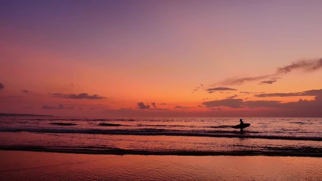 Colorful vivid sunset sky and surfer sulhouette carrying surfboard on tropical sea beach of Bali island. Slow motion video of beautiful tropical nature in Indonesia