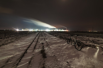 A night view of an industrial unit, a wood-processing plant with fuming pipes and a high-illuminated area on the background of a snow-covered field and a bicycle with a red light.