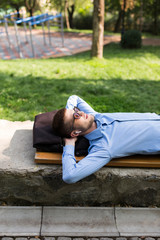Obraz premium Young attractive man in blue shirt and sunglasses with wireless earphones dreamily lying on bench in park