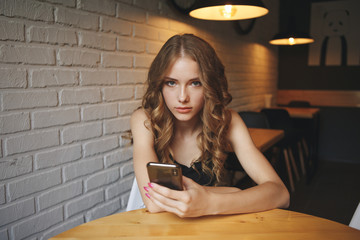 Sad young girl sitting in a cafe looking tired her phone, unhappy girl watching her black phone, tiered blonde girl reading a bully message online