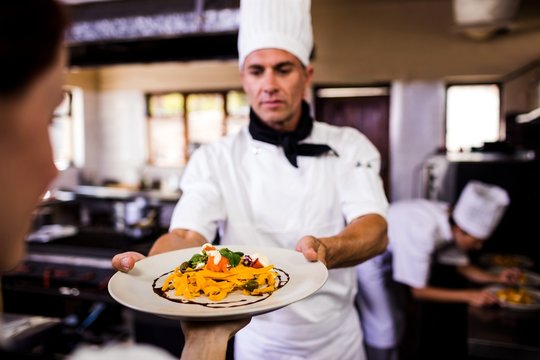 Male Chef Giving Plate Of Prepared Food To Waitress In Kitchen