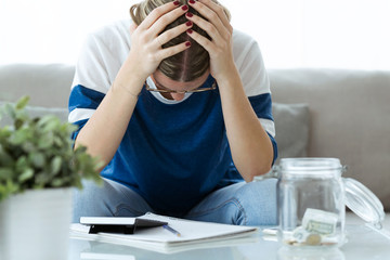 Depressed young woman holding her head with hands while counting savings at home.