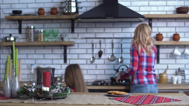 Cute mother in plaid shirt with long blond hair cooking breakfast and small naughty girl bite one cookie