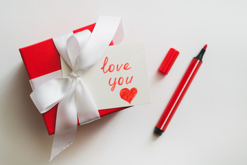 Red gift boxes tied with a white ribbon, a marker and a card with an inscription "love you" on a light background. The process of preparing a gift for a loved one.