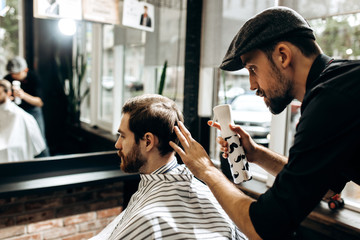 Mustachioed barber dressed in a black shirt is doing the hair styling to the stylish man sitting in...