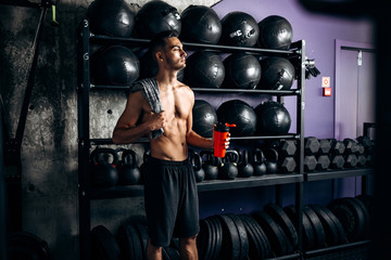 Fototapeta na wymiar Athletic dark-haired man with a naked torso dressed in the black shorts is standing near the sport equipment in the gym and holds bottle with water in the hand