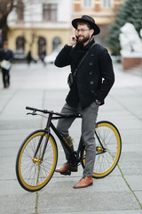 Inviting friend for a ride. Happy young bearded man talking on the mobile phone and smiling while sitting near his bicycle outdoors